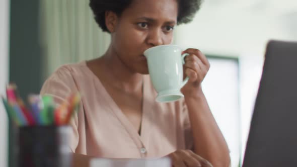 Happy african american woman sitting at table using laptop and drinking coffee