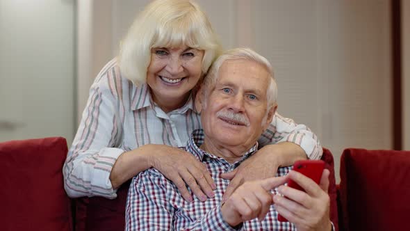 Happy Smiling Senior Couple with Mobile Phone at Home. Resting on Sofa in Cozy Living Room