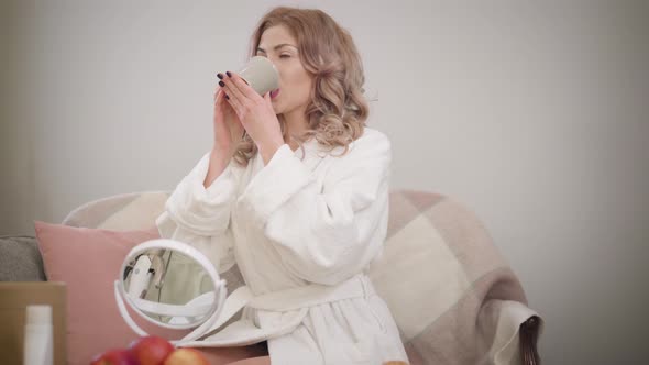 Portrait of Happy Caucasian Girl in White Bathrobe Drinking Coffee. Charming Confident Woman with