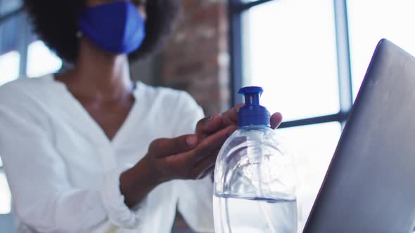 African american woman wearing face mask sitting in cafe with laptop disinfecting hands