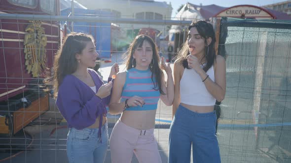 Three Happy Girls with Lollipops Stand and Joke Around By Metal Fence