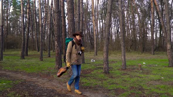 A Backpacker with a Guitar and a Camera Walks Along a Trail in the Forest