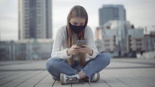 Portrait of Concentrated Young Woman in Covid Face Mask Sitting on City Square and Messaging Online