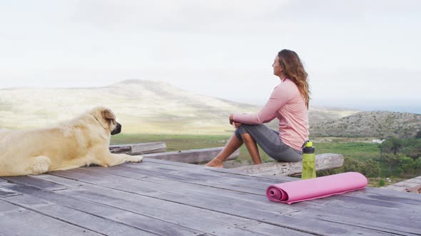 Caucasian woman sitting outdoors on deck with pet dog admiring view in rural mountainside setting