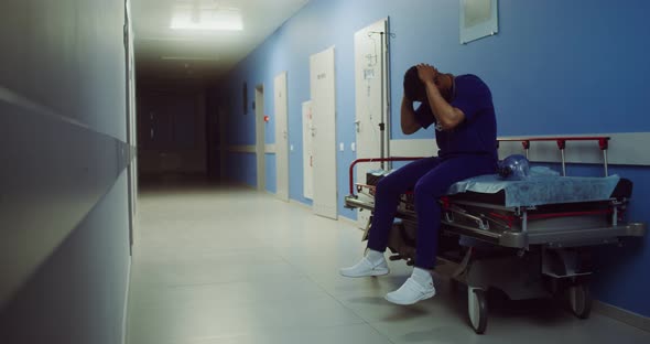 A Doctor Sits on a Medical Gurney and Rubs His Head in Empty Hospital Corridor