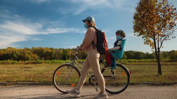 Mom with a Child in Medical Masks Walk with a Bicycle