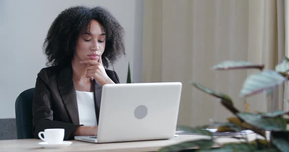 Pensive Girl Sits at Home at Table with Laptop, Processes Information, Frowns Serious Young Face