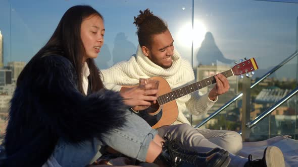 Talented African American Young Man Playing Guitar As Asian Woman Singing Sitting on Rooftop in