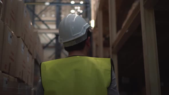 Young Warehouse Worker with Safety Hard Hat is Walking Through Inventory Room