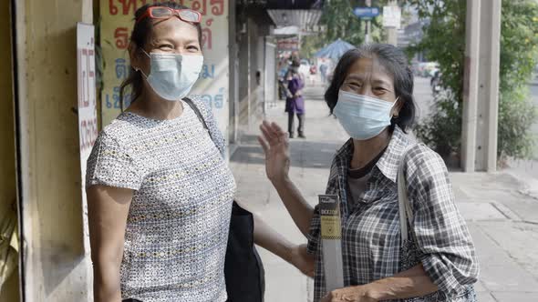 Two Happy Adult Thai Women Wearing Facemask Smiling Waving To The Camera At The Street Sidewalk