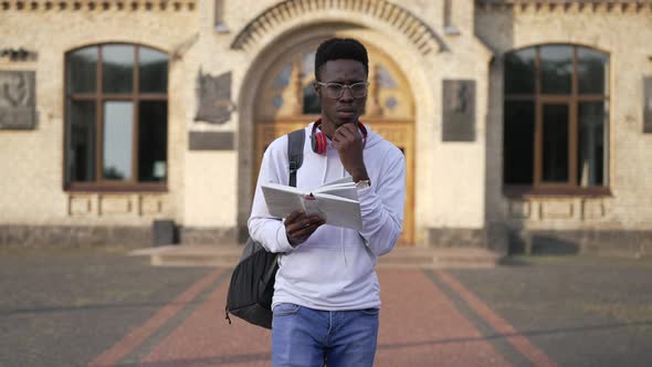 Thoughtful Smart African American Young Man Walking at University Campus Reading Book