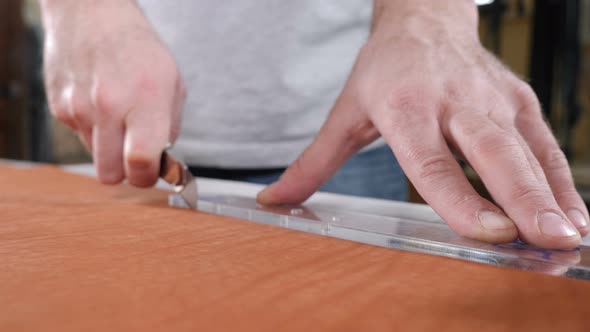 Hands of Skilled Leather Craftsman Using Plastic Pattern and Leather Knife to Cut Out Part From