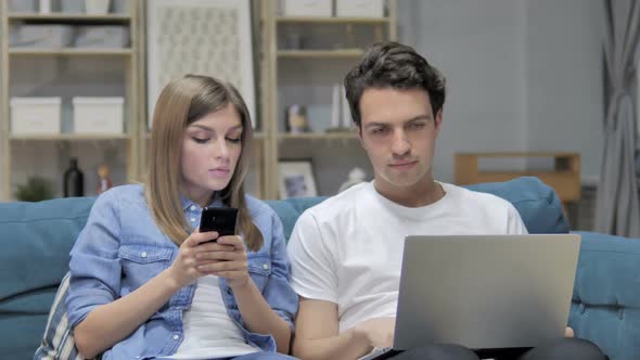 Young Couple Talking While Using Smartphone and Laptop at Home