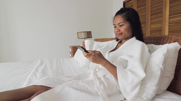 Adult woman drinking coffee and using her mobile phone, browsin the internet while laying on a white