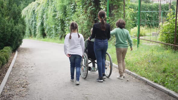 Back View Caucasian Children Pushing Wheelchair with Disabled Friend Outdoors Walking in Slow Motion