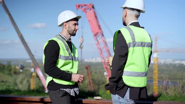 Side View Two Young Positive Men Talking Standing at Lunch Break on Construction Site Outdoors