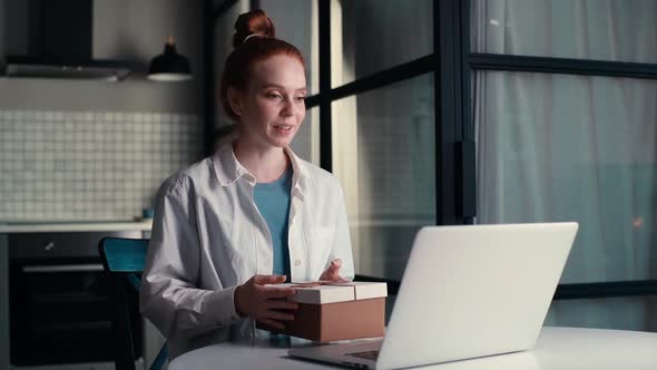 Portrait of Charming Young Woman Opening Gift Box with Present While Having Video Call on Laptop