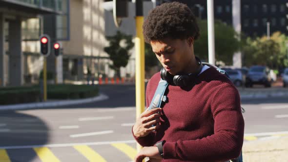 African american man in city checking smartwatch, wearing headphones and backpack waiting in street