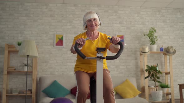 Active Old Woman in Headphones Engaged in Cardio Training on a Exercise Bike