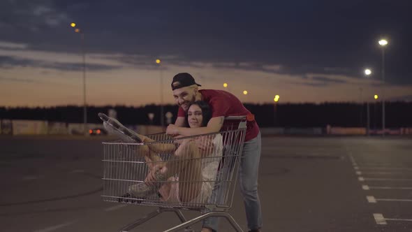 Camera Approaches To Happy Young Man Hugging Woman Sitting in Shopping Cart. Portrait of Cheerful