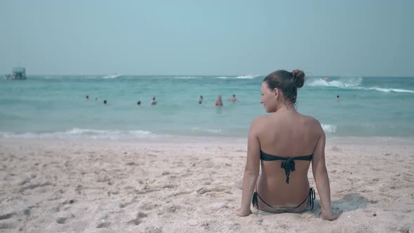 Brunette in Black Bikini Sits on Yellow Sand Ocean Beach