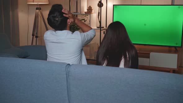 Back View Of Upset Young Asian Watching Tv With Mock Up Green Screen In The Living Room At Home