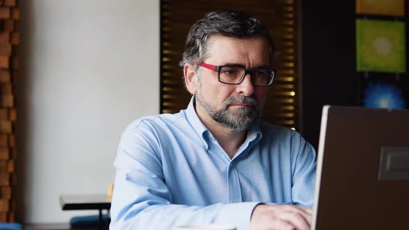 Side View of Senior Man Who Sits in Cafe in Daytime and Using the Laptop for the Remote Work