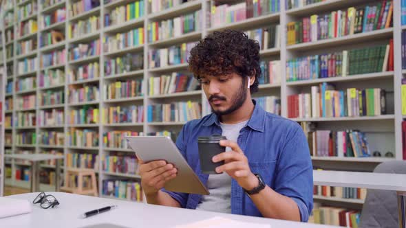 Young Indian Male Student Wearing Earphones Use Mobile Tablet Sitting in Library