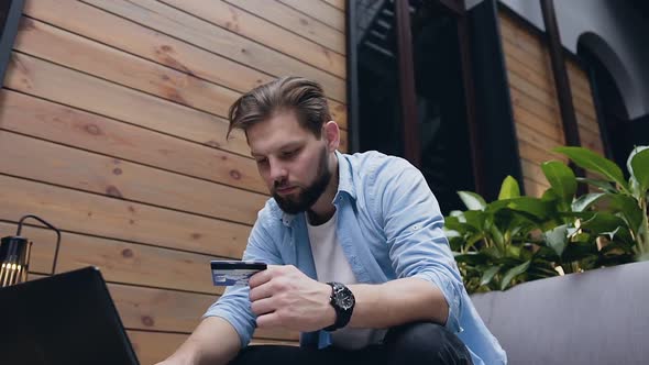 Bearded Young Man which Entering Credit Card Details on Keyboard while Sitting in Hall