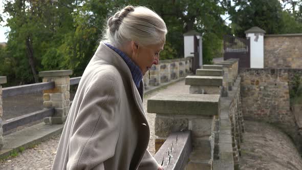 An Elderly Woman Leans Against a Wooden Railing and Looks Around Throughtfully