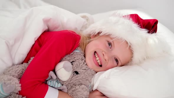 Smiling Girl in Santa Hat and Christmas Pajamas Lying in Bed with Teddy Bear at Home