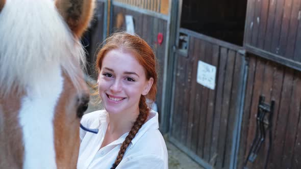 Young female rider standing by horse in stable, smiling