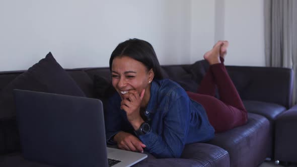 Mixed race woman lying on couch using laptop and laughing