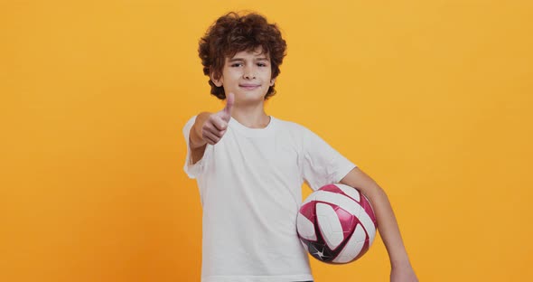 Little Football Player Standing with Soccer Ball and Gesturing Thumb Up, Orange Studio Background