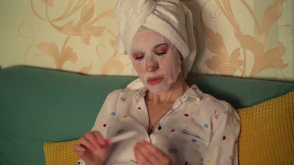 Portrait, a Woman in a Cosmetic Fabric Mask and with a White Towel on Her Head, Files Her Nails with