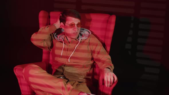 Thoughtful Man in Glasses Sits in Armchair in Dark Room with Falling Red Light and Shadow Blinds