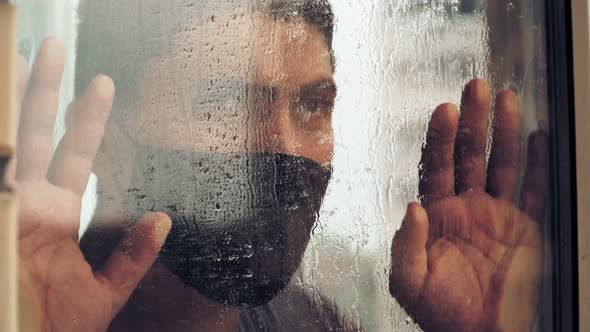 Sad man in medical mask walk window wet after rain to look outside during quarantine due coronavirus