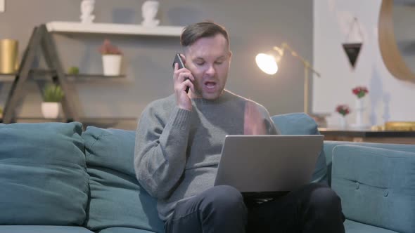 Angry Casual Man with Laptop Talking on Smartphone on Sofa
