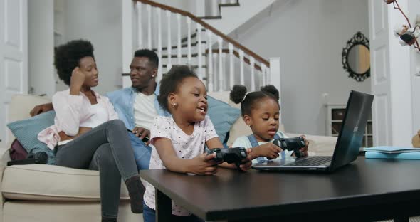 African American Couple Sitting on Soft Couch while Their Two Small Daughters Gaming on Laptop