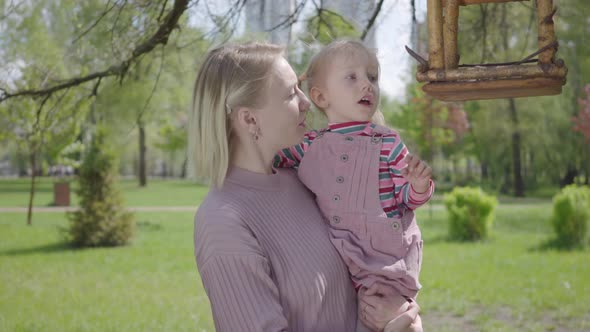 Portrait Young Blond Mother Hold Adorable Small Daughter and Show Her Bird Feeder House in the