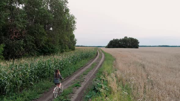 Aerial Shot of Young Girl Riding Bike Through Meadow Fields on Country Road at Sunset