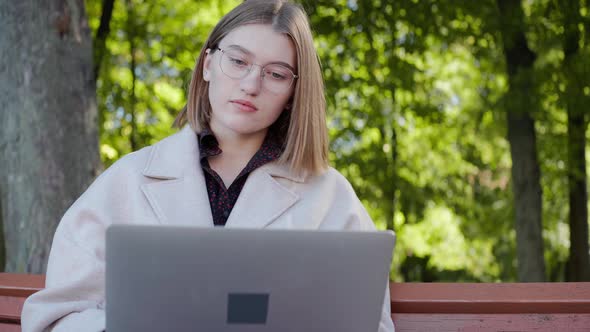 Girl Sits on a Bench in Autumn Writing on a Laptop and Looking Around Smiling