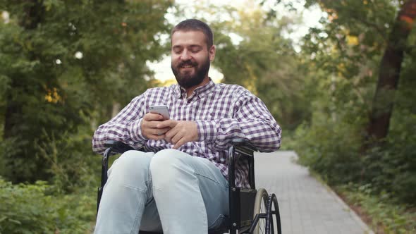 Emotional Hipster Disabled Man Sits on Wheelchair in Park and Uses Phone