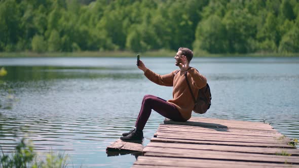 A Man with a Backpack Sits on the Dock of the Lake and Calls By Video Link on a Mobile Phone