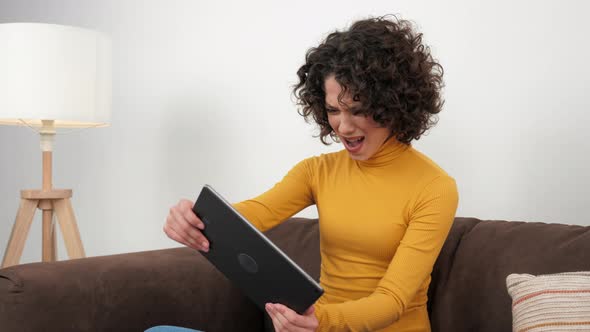 Curly Woman Emotionally Playing Video Game on Tablet Sitting on Couch at Home