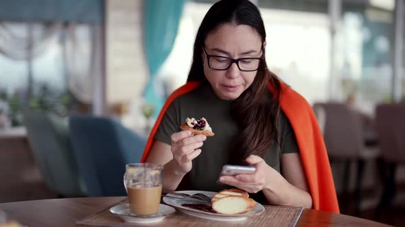 Woman is Having Breakfast in Restaurant Eating Cake and Coffee Surfing Internet By Smartphone