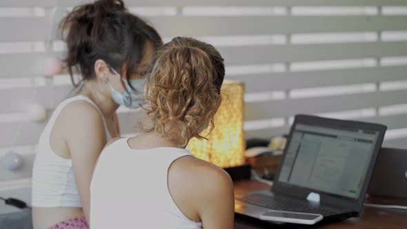 Two Young Women Wearing a Protective Mask Sitting at Table in Cafe with Laptop Works Together