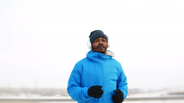 Black Man Wearing Headphones and Winter Sportswear Trains Outdoors While Jogging Front View