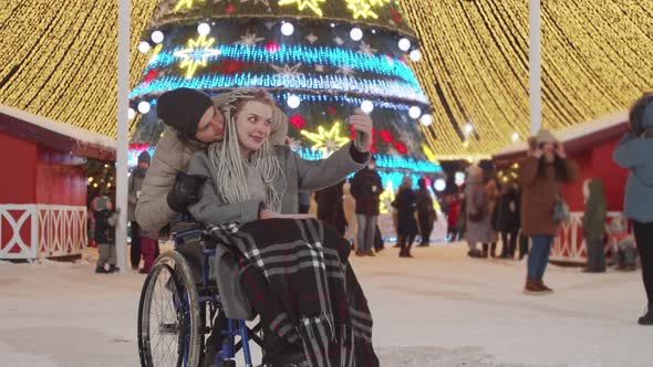 Smiling Young Woman in a Wheelchair and Her Boyfriend at Christmas Party on the Streets Taking a