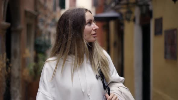 Attractive Woman Walks Along a City Narrow Street with Backpack Looks with Interest Around